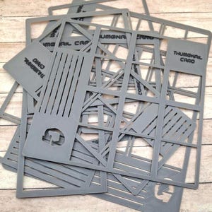 Puede incluir: Un conjunto de plantillas de plástico gris con varios recortes geométricos y lineales. Las plantillas tienen la inscripción "THUMBNAIL CARD". Las plantillas están dispersas sobre una superficie de madera clara.