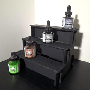 May include: A black tiered display stand holding four bottles of Dr. Ph. Martin's Bombay India Ink in various colors. The bottles have dropper tops and labels with the brand name and ink color.
