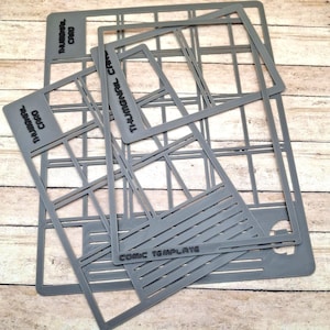 May include: A collection of gray plastic stencils for crafting. The stencils include rectangular and grid-like shapes, with text that reads "THUMBNAIL CARD" and "COMIC TEMPLATE". The stencils are arranged on a light-colored wooden surface.