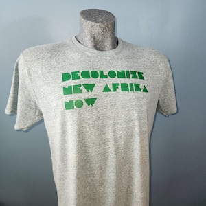 May include: A gray t-shirt with the text "DECOLONIZE NEW AFRIKA NOW" printed in green.