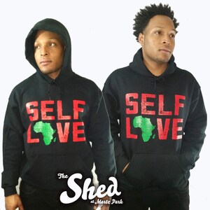 May include: Two people wearing black hoodies with the words "SELF LOVE" in red and green on the front. The green is in the shape of the continent of Africa. The hoodies also have the words "The Shed at Martz Park" on the bottom.