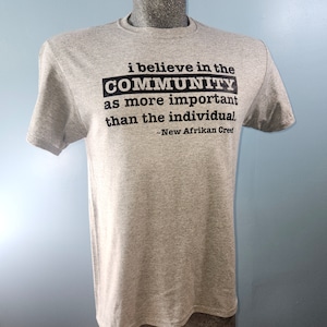 May include: Gray t-shirt with black text that reads "i believe in the COMMUNITY as more important than the individual, -New Afrikan Cred"