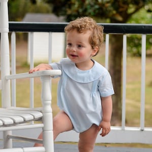 May include: A young child wearing a light blue short-sleeved onesie with a Peter Pan collar and a ruffled bib. The child is standing on a white rocking chair with a wooden frame.