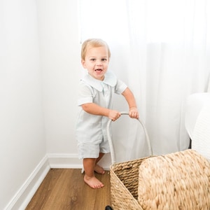 May include: A young child in a light blue collared shirt and matching shorts pushes a wicker basket on wheels. The basket is filled with a woven cushion.