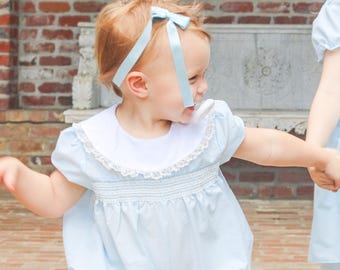 May include: A young child wearing a light blue romper with a white collar and lace trim. The romper has a gathered waist and short sleeves. A light blue bow is in the child's hair. The background features a brick wall.
