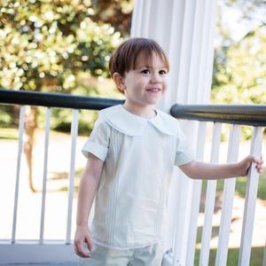 May include: A young boy wearing a light green short-sleeved button-down shirt with a white collar and matching shorts. He is standing on a porch and smiling.