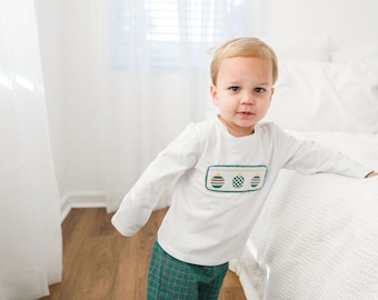 May include: A young child wearing a white long-sleeved shirt with a green and red embroidered ornament design and green and red plaid pants. The child is standing in front of a white bed.