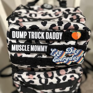 Pu&ograve; includere: Uno zaino a stampa leopardata bianco e nero con spalline nere e pi&ugrave; tasche. Lo zaino ha tre toppe attaccate sul davanti: una toppa nera con testo bianco che dice "DUMP TRUCK DADDY", una toppa nera con testo bianco che dice "MUSCLE MOMMY" e una toppa colorata con il testo "Leg Day Everyday".