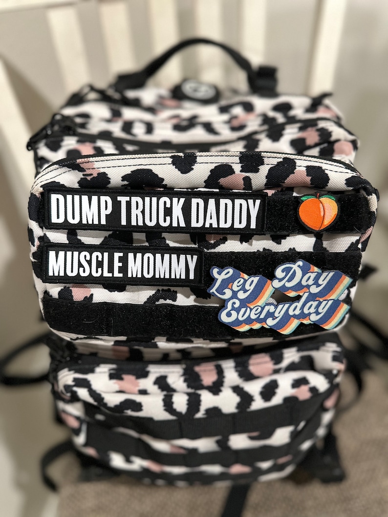 Pu&ograve; includere: Uno zaino a stampa leopardata bianco e nero con spalline nere e pi&ugrave; tasche. Lo zaino ha tre toppe attaccate sul davanti: una toppa nera con testo bianco che dice "DUMP TRUCK DADDY", una toppa nera con testo bianco che dice "MUSCLE MOMMY" e una toppa colorata con il testo "Leg Day Everyday".