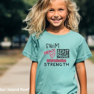 May include: A teal blue t-shirt with the text "Swim Heart Beast Mode Determination Strength" printed on the front. The text is in black with pink accents.