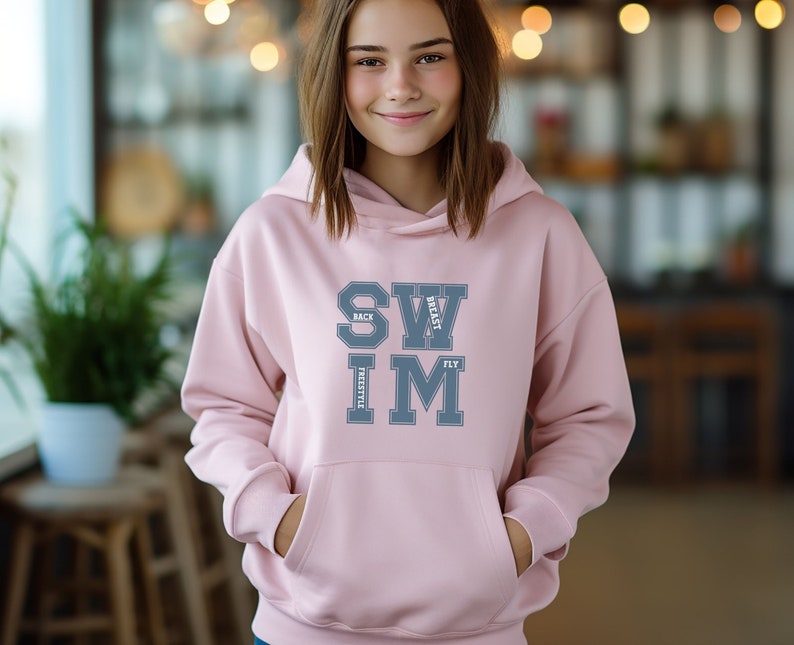 May include: A light pink hoodie with a front pocket and a drawstring hood. The word "SWIM" is printed in gray block letters, with "BACK", "BREAST", "FREESTYLE", and "FLY" arranged around it. The hoodie is made of a soft material.