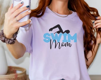 May include: Orchid-colored t-shirt with the text "Swim Mom" in blue and black. The design includes a swimmer graphic. The person in the image is holding a phone.