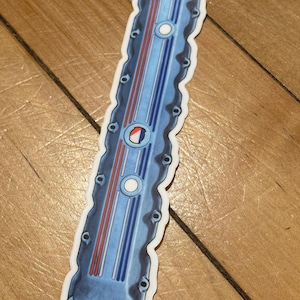 Puede incluir: Una pegatina con una tapa de válvula de motor azul con rayas rojas y blancas. La pegatina tiene un borde blanco y está sobre una superficie de madera. La tapa de la válvula tiene un emblema circular y varios orificios para pernos.