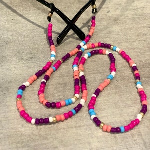 Può includere: Una catena per occhiali con perline colorate, con perline rosa, viola, bianche e blu. La catena è attaccata a un paio di occhiali da sole neri.