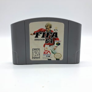 May include: A gray Nintendo 64 game cartridge for FIFA Soccer 64. The front of the cartridge features a soccer player in a red and white uniform, a soccer ball, and the game title.