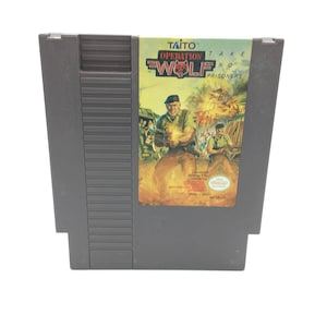 Op de afbeelding: Een grijze Nintendo Entertainment System (NES) spelcartridge voor "Operation Wolf". Het label toont een soldaat met een wapen, met de titel en het logo van het spel in vette letters. De cartridge is rechthoekig.