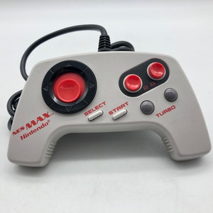 Könnte beinhalten: Ein weißer NES MAX-Controller mit roten Knöpfen und einem schwarzen Steuerkreuz. Der Controller hat einen grauen Turbo-Knopf und den Text "NES MAX" und "Nintendo" auf der Seite.