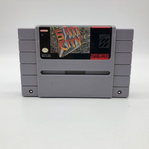 May include: A gray Super Nintendo Entertainment System game cartridge with a black label. The label features a city skyline and the text "SimCity" in red and yellow. The text "Super Nintendo" is printed in white on a black background.