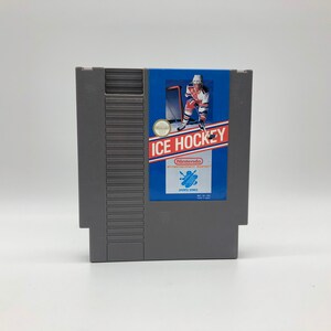 May include: A gray Nintendo Entertainment System (NES) game cartridge for Ice Hockey. The front of the cartridge features a blue and white label with a hockey player in a red, white, and blue uniform skating with a hockey stick. The label also includes the text "ICE HOCKEY" and the Nintendo logo.