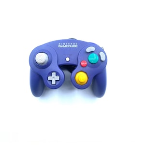 Może przedstawiać: Fioletowy kontroler Nintendo GameCube z szarym analogowym joystickiem, żółtym joystickiem C i zielonymi i czerwonymi przyciskami. Kontroler ma napis "NINTENDO GAMECUBE" nadrukowany z przodu.