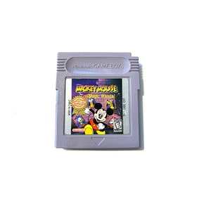May include: A gray Game Boy Color cartridge with a purple label featuring Mickey Mouse and the text "Mickey Mouse Magic Wands!" The label also includes a Nintendo Seal of Quality and an ESRB rating of "E" for Everyone.