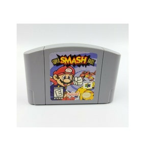 Puede incluir: Un cartucho de juego gris de Nintendo 64 para Super Smash Bros. La parte frontal del cartucho presenta una imagen colorida de Mario golpeando a un personaje rojo y amarillo. El juego está clasificado como E para todos por la ESRB.