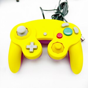 May include: A yellow and purple GameCube controller with a gray analog stick and buttons. The controller has a black cord attached to it.