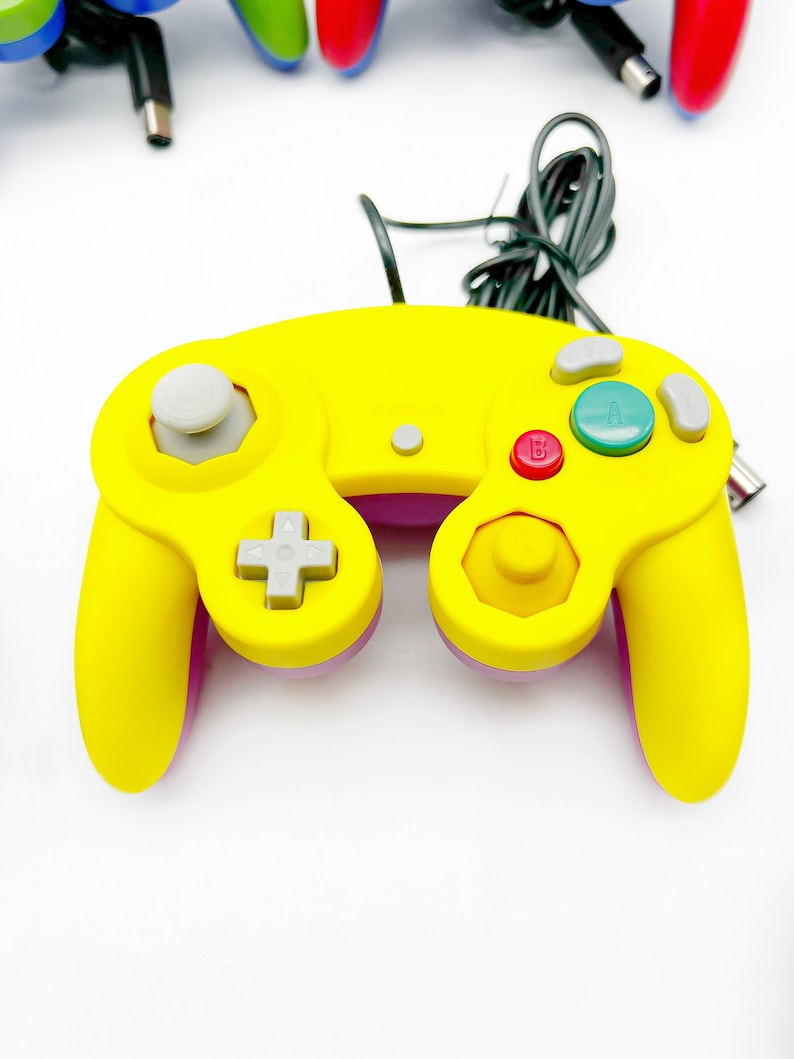 May include: A yellow and purple GameCube controller with a gray analog stick and buttons. The controller has a black cord attached to it.