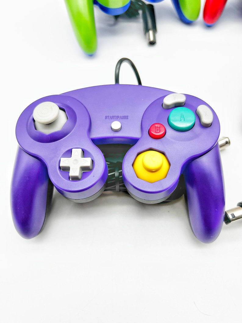 May include: A purple GameCube controller with a yellow, red, and green button. The controller has a white plus sign button and a white start/pause button.