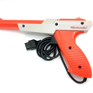 May include: A red and white Nintendo Zapper light gun for the NES video game console. The gun has a black cord and a black connector. The gun has the word "Nintendo" printed on the side.