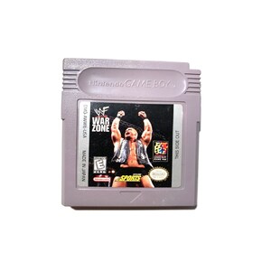 May include: A gray Game Boy Color cartridge with a black label. The label features a wrestler in a black and white wrestling outfit, with the text "WWF War Zone" and "This Side Out".