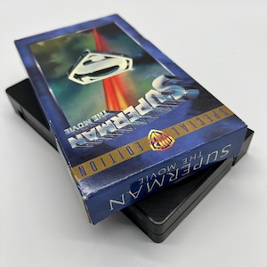 Könnte beinhalten: Ein VHS-Band des Films "Superman: The Movie" in einem blauen und goldenen Etui mit einem Special Edition-Label. Das Etui zeigt ein Bild von Superman, der mit einem roten und orangefarbenen Laserstrahl hinter sich durch die Luft fliegt.