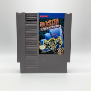 Könnte beinhalten: Eine graue Nintendo Entertainment System (NES) Spielkassette mit einem Etikett, auf dem "SUNSOFT" und "BLASTER MASTER" steht. Das Etikett zeigt einen blauen und schwarzen Hintergrund mit einem roten Ziel und einem grünen Monster.