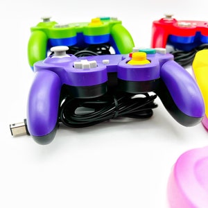 May include: A purple GameCube controller with a black cord and yellow, blue, and gray buttons. The controller is on a white surface.