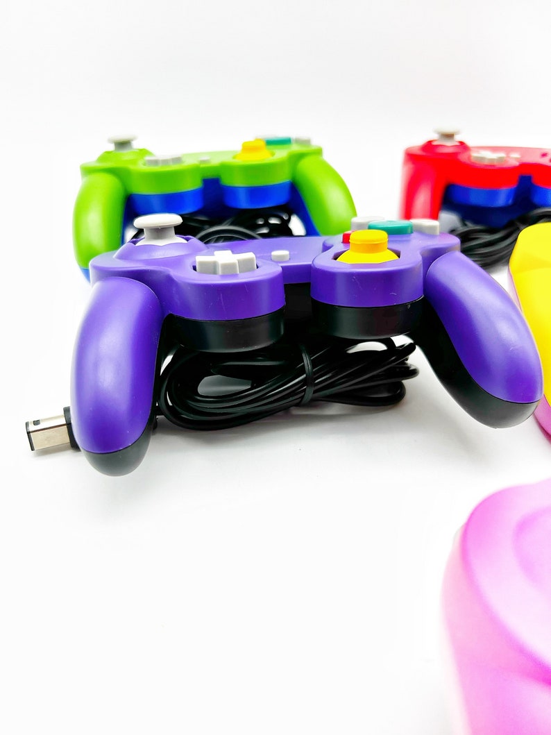 May include: A purple GameCube controller with a black cord and yellow, blue, and gray buttons. The controller is on a white surface.