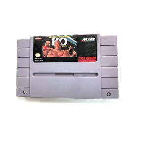 May include: A gray Super Nintendo Entertainment System (SNES) game cartridge with a label featuring a boxing match. The label reads "George Foreman's KO Boxing" and "Acclaim Entertainment".