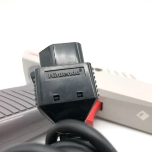 May include: Close-up of a black Nintendo controller connector with the word "Nintendo" printed on it. The connector is attached to a gray and white cord.
