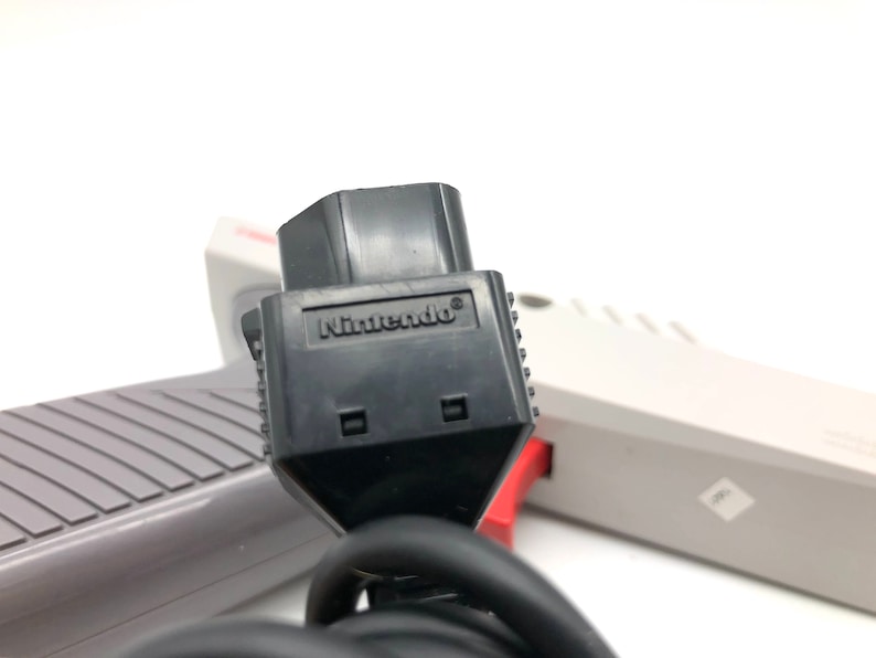 May include: Close-up of a black Nintendo controller connector with the word "Nintendo" printed on it. The connector is attached to a gray and white cord.