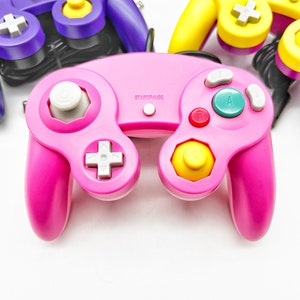 May include: A pink Nintendo GameCube controller with white buttons and a yellow C-stick. The controller has a START/PAUSE button and the letters A and B on the face buttons.