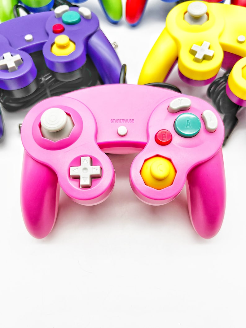 May include: A pink Nintendo GameCube controller with white buttons and a yellow C-stick. The controller has a START/PAUSE button and the letters A and B on the face buttons.