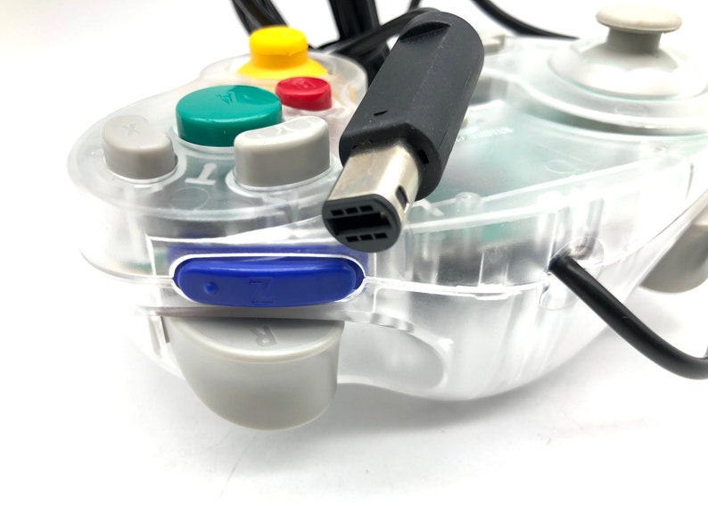 May include: A clear, transparent GameCube controller with a black cable and a blue button. The controller has a green, yellow, and red button.