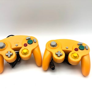 Pode incluir: Dois controladores amarelos do GameCube com botões cinzentos e verdes. Os controladores estão em uma superfície branca.