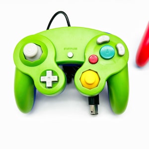 May include: A green GameCube controller with a yellow, red, and blue button layout. The controller has a black cord and the text "START/PAUSE" is printed on the top.
