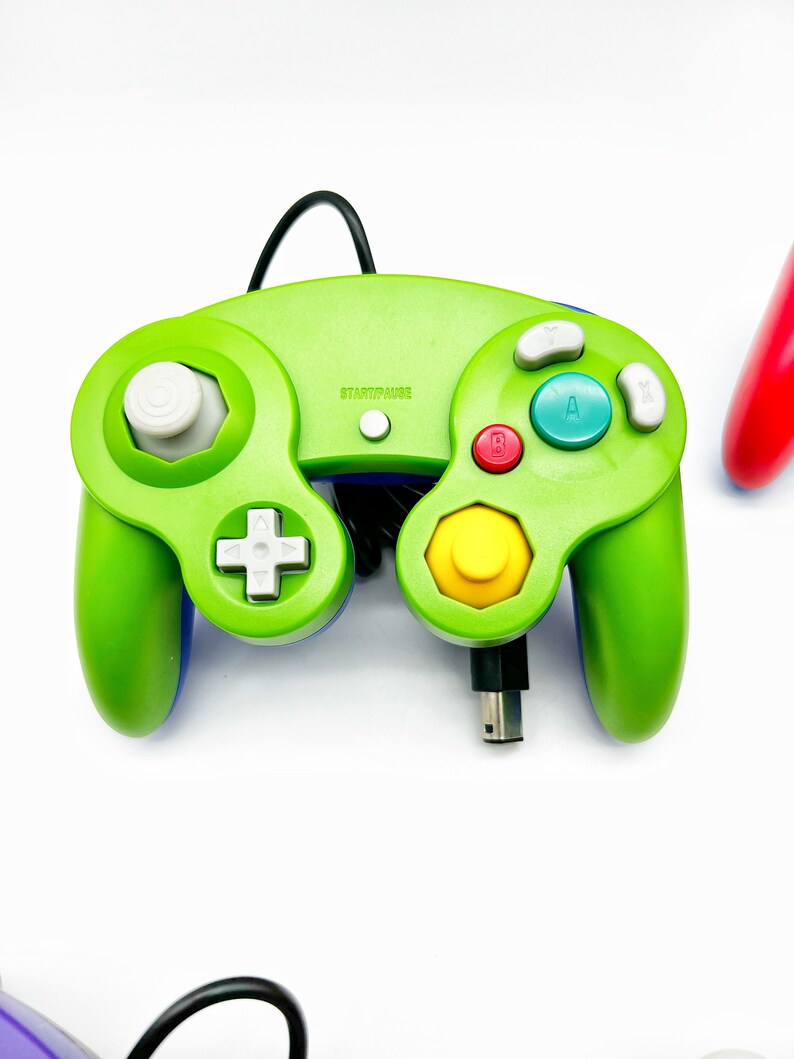 May include: A green GameCube controller with a yellow, red, and blue button layout. The controller has a black cord and the text "START/PAUSE" is printed on the top.