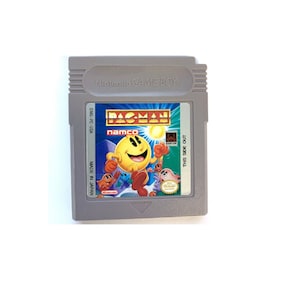 Könnte beinhalten: Eine graue Game Boy Color-Cartridge mit einem blauen und gelben Etikett. Das Etikett zeigt den Text "PAC-MAN" in einem weißen Feld mit gelbem Rand. Der Text "namco" befindet sich unter dem Titel in einem weißen Feld mit blauem Rand. Das Etikett zeigt auch ein Cartoon-Bild von Pac-Man, der mit Geistern rennt.