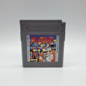 Könnte beinhalten: Eine graue Game Boy-Cartridge mit einem Etikett, auf dem "Dr. Mario" und "Nintendo Game Boy" steht. Das Etikett zeigt auch ein Bild von Mario, der als Arzt verkleidet ist.