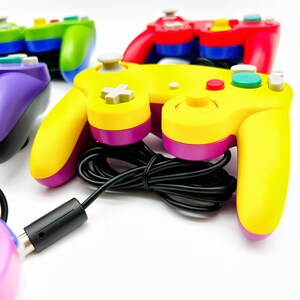 May include: A yellow and purple GameCube controller with a black cord. The controller has a gray button in the center and a red button on the right side.