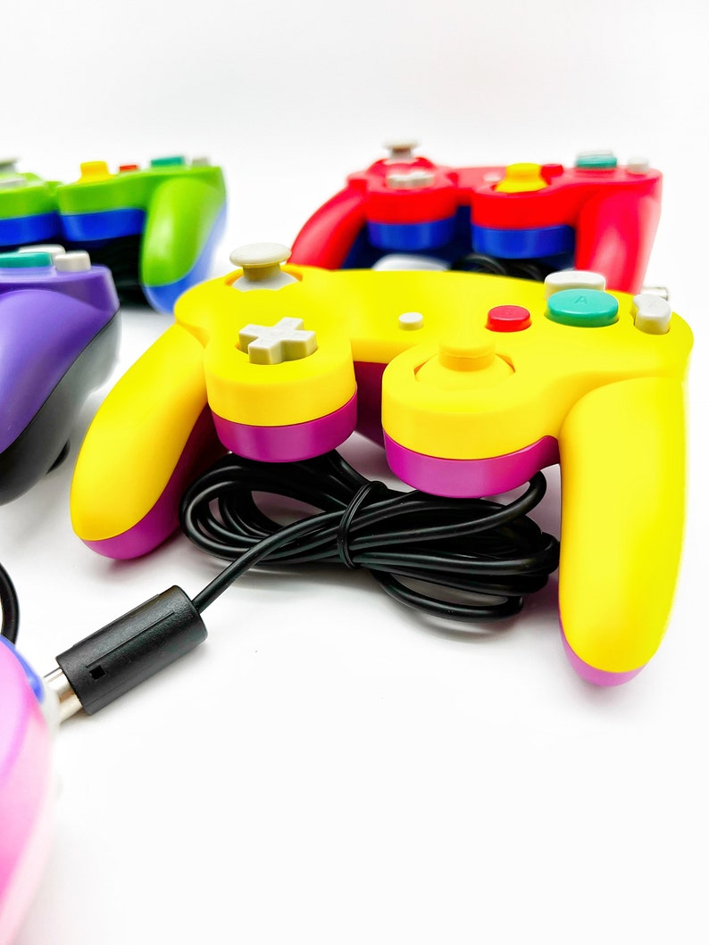May include: A yellow and purple GameCube controller with a black cord. The controller has a gray button in the center and a red button on the right side.