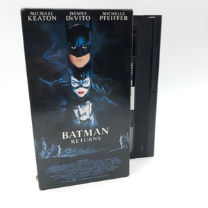 May include: A VHS tape case for the movie Batman Returns. The case features a black background with a photo of Batman and Catwoman. The title of the movie is in white text. The actors' names are listed on the front of the case: Michael Keaton, Danny DeVito, and Michelle Pfeiffer.
