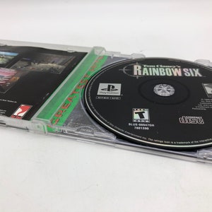 Puede incluir: Una caja de juego de PlayStation para Tom Clancy's Rainbow Six. La caja es negra con un borde verde y blanco. El disco del juego es visible dentro de la caja. El juego est&aacute; clasificado como T para adolescentes por la ESRB.